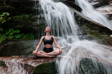 Young woman sitting in lotus pose under tropical waterfall meditating with calm strength and mindfulness fully connected to nature energy tranquility and inner balance