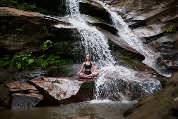 Young woman sitting in lotus pose under tropical waterfall meditating with calm strength and mindfulness fully connected to nature energy tranquility and inner balance