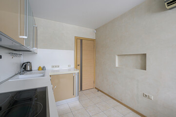 bright kitchen corner features beige cabinets, a sink, and a cooktop. A door and a recessed niche are visible in the textured wall