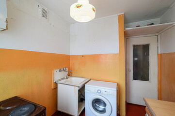 small, dated kitchen with two-tone walls, a sink, a washing machine, and a door. The room has a...