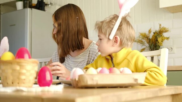 Children siblings painting Easter egg sitting together at table. Easter Family traditions. Teen girl teaching happy little brother and sister to dye, decorate eggs with paints. Preparation to holidays