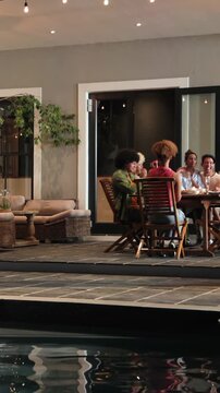 Vertical video: Diverse friends greeting at table, raising flutes for toast under string lights