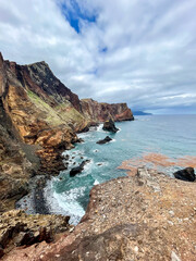 A dramatic view of the rugged cliffs and rocky shoreline near Caniçal, Madeira. The ocean waves crash against the volcanic rocks.