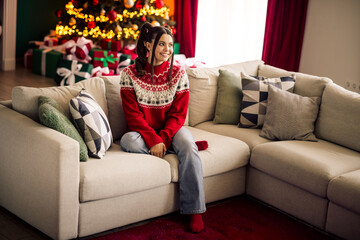 Obraz premium Young woman enjoying festive moments at home in a Christmas sweater, surrounded by holiday decorations and a lit tree.