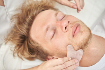 Young caucasian male relaxing with facial gua sha massage for skin rejuvenation and stress relief.