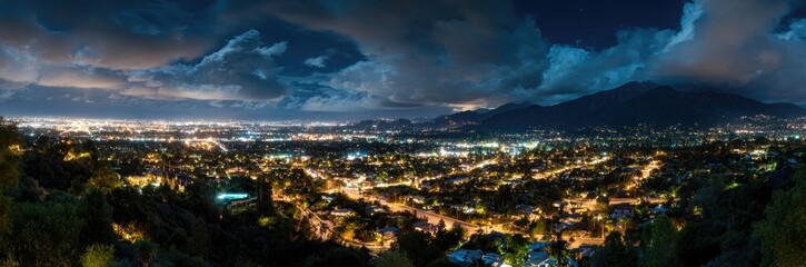 Fototapeta premium Night cityscape panorama, with illuminated city lights nestled between hills and mountains under a dramatic sky