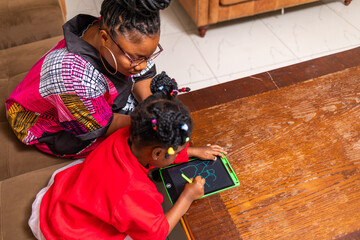 Early education, African mother watching her daughter write alphabets on a digital tablet. Parent and child early literacy at home