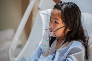Sick Asian girl wearing oxygen mask lying on hospital bed receiving treatment. Healthcare concept, respiratory disease, hospital admission, medical care, and child patient in emergency situation.