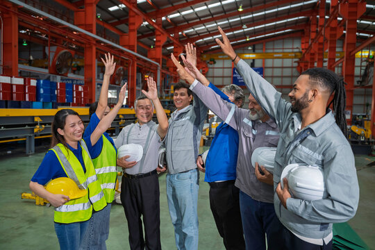 Happy multicultural industrial workers celebrating success with high fives inside a factory warehouse, showing motivation, teamwork, diversity, and positive energy in a professional workplace.