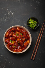 Stir fried rice cakes tteokbokki simmered in spicy sauce, garnished with green onions and sesame seeds, served on a black background with chopsticks top view. Popular dish of Korea