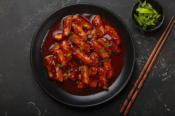 Stir fried rice cakes tteokbokki simmered in spicy sauce, garnished with green onions and sesame seeds, served on a black background with chopsticks top view. Popular dish of Korea