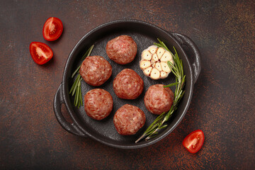 Raw meatballs seasoned with garlic and rosemary ready to be cooked in a cast iron pan, on a dark brown table with a red checkered napkin and cherry tomatoes