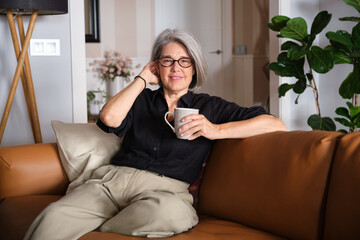 Smiling mature woman relaxing on comfortable sofa holding a mug and enjoying a cup of coffee or tea at home