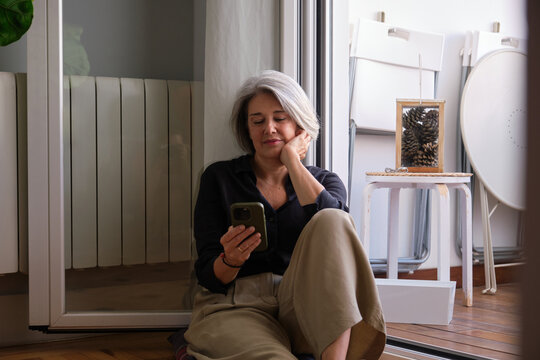 Relaxed mature woman using mobile phone app sitting on floor next to balcony door at home - Powered by Adobe