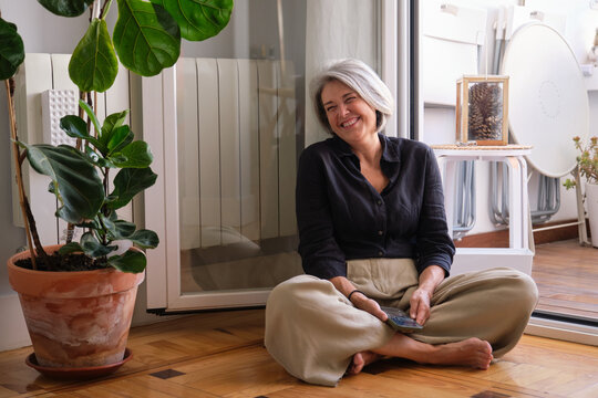 Happy mature woman sitting on the floor with legs crossed holding a smartphone, laughing