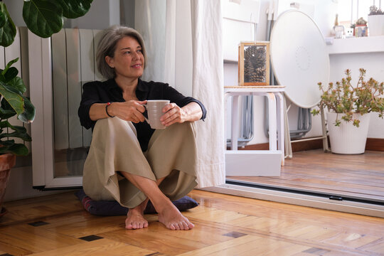 Serene mature woman enjoying a quiet moment with a cup of coffee, sitting on the floor by her balcony door, embracing the tranquility of her home