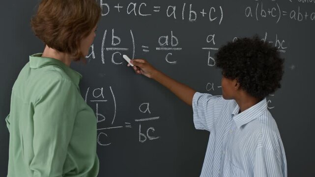 African American student using chalk to point out fraction-based algebraic equation on blackboard, guided by observing teacher