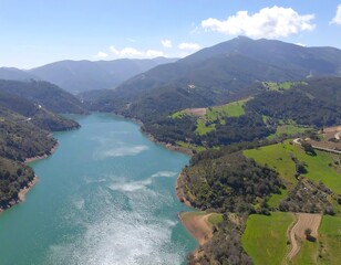 High-angle view of a turquoise lake, winding through valleys, nestled amidst lush green hills and mountains, under a partly cloudy sky
