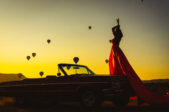Silhouette of a Woman in Red Dress and Vintage Convertible Car at Sunrise with Hot Air Balloons in Cappadocia