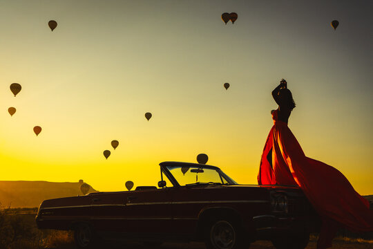 Silhouette of a Woman in Red Dress and Vintage Convertible Car at Sunrise with Hot Air Balloons in Cappadocia