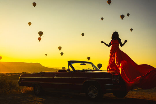 Silhouette of a Woman in Red Dress and Vintage Convertible Car at Sunrise with Hot Air Balloons in Cappadocia