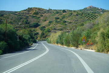A scenic view of a winding asphalt road, part of the Northern Road Axis of Crete (BOAK), curving through the arid, scrub-covered hills of the Greek island under a bright blue sky.