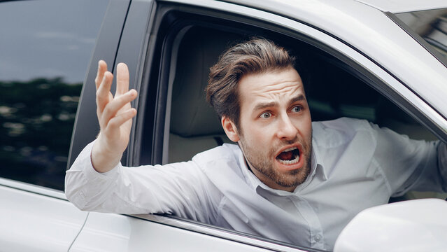 Angry driver stuck in traffic jam gesticulates with hand and screaming. Crazy man yelling from car. Stress, traffic rush hour. Angry aggressive guy driving in auto shouting up through open window