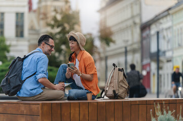 Smiling tourists with backpacks sitting outdoors, reading a map and using a smartphone while planning their city sightseeing adventure during vacation