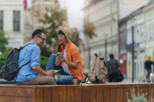 Smiling tourists with backpacks sitting outdoors, reading a map and using a smartphone while planning their city sightseeing adventure during vacation