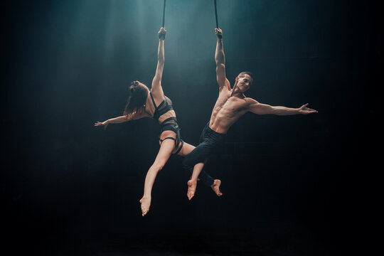 Duo aerial straps performance with male and female acrobats. Both extend arms outward in mirrored pose, suspended mid-air under dramatic blue stage spotlight.