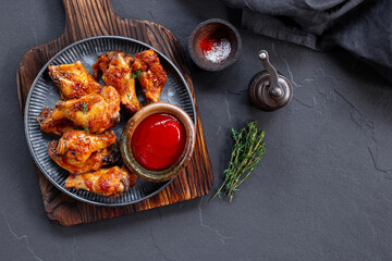 Grilled spicy chicken wings with ketchup in a plate on a dark slate, stone or concrete background. Top view with copy space.
