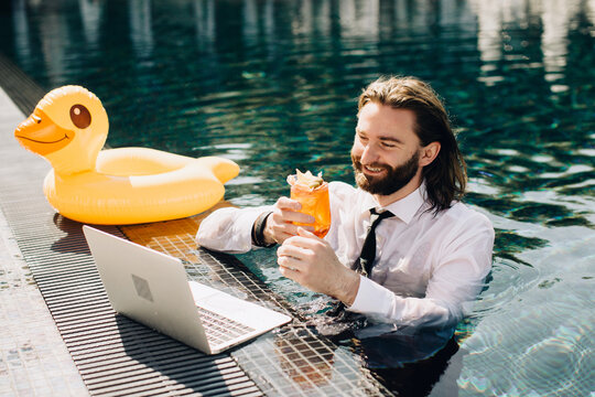 Businessman working remotely from swimming pool with laptop and cocktail