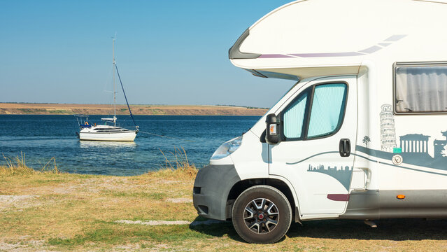 panoramic view to family motorhome on the coast and white sail boat in bay under blue sky
