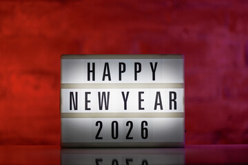 A cinematic lightbox with the words Happy New Year 2026 illuminated, placed against a rustic brick background on a glossy white reflective table