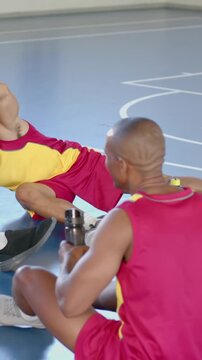 Sitting diverse male teammates holding water bottle and stretching leg on gym floor, with markings
