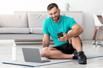 Happy smiling young caucasian guy sitting on mat on floor typing on phone and looking at computer in living room interior. Covid-19 quarantine and video call, communication remotely and trainer online