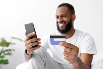Happy young african american guy with beard with smartphone shopping online with credit card in bedroom interior. Buying purchases at home, store remotely, banking and paying for goods, sale and offer
