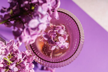 Closeup of a textured champagne glass with sparkling wine and lilac petals on a vibrant violet background