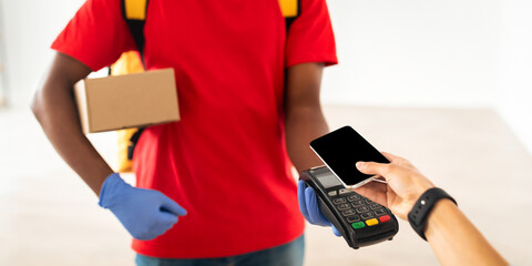Pov point of view of customer using smartphone with blank empty screen for contactless payment, black male courier in protective gloves holding Point Of Sale machine and cardboard box with purchase