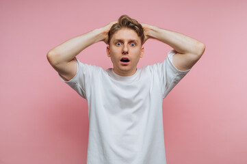 Young man shows surprise while holding his head in a playful manner against a pink background in a...