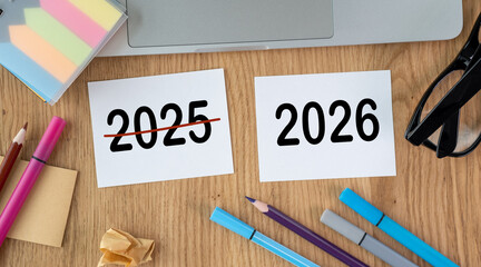 Paper with crossed out inscription 2025 and 2026 on wooden desktop. Planning plans for New Year 2026. Laptop, glasses, pen.