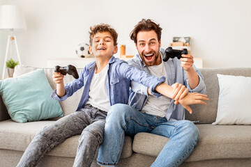 Joyful dad and son with joysticks playing video games at home, boy distracting father with hand, faving fun together, sitting on sofa in living room. Fatherhood and leisure activities concept