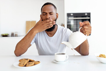 Lack Of Sleep Concept. Portrait of tired young African American man yawning while sitting at dining table in kitchen, holding teapot pouring coffee away from cup spilling hot drink on desk