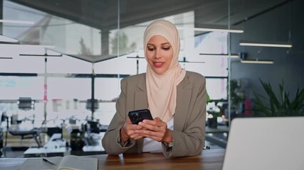 Middle Eastern muslim businesswoman manager ceo using cell phone mobile app. Smiling young indian woman in hijab holding smartphone sitting in office working online at desk on gadget with copy space. - Powered by Adobe