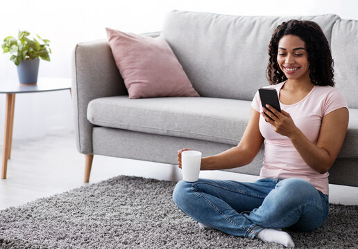 Video call conference, social distancing. Happy young black lady have chat on mobile phone at home, talk on smartphone, female greeting with family, friend online, hold cup of drink in living room
