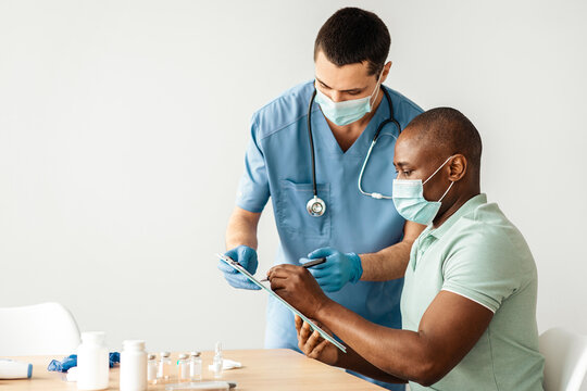 Preparation for coronavirus immunization and health care during pandemic. Young doctor in protective mask and adult african american patient fill out questionnaire for procedure at table, empty space