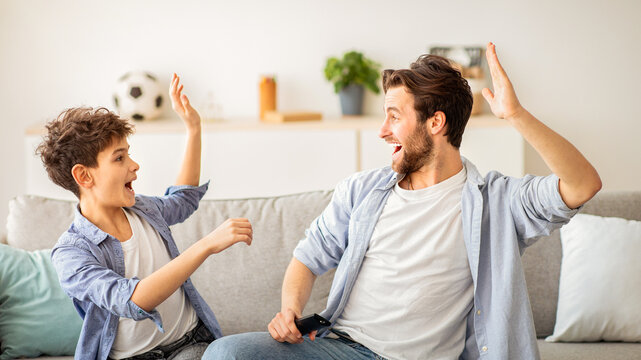 Happy young father and teen son giving high-five while watching soccer match and cheering for favourite team, sitting together on sofa in living room. Fatherhood, single parent concept