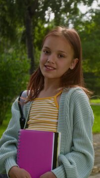 Vertical portrait shot of teenage girl wearing striped shirt and cardigan, smiling at camera while holding pink notebook outdoors