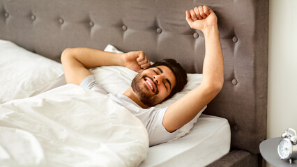 Rested arab man waking up and stretching hands, wellslept guy lying in bed, awakening in cozy bedroom and smiling with eyes closed. Good rest, sweet dreams and recreation concept