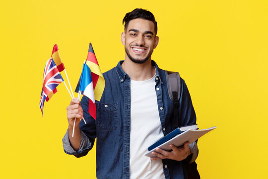 Positive young middle eastern man in casual with backpack and bunch of books student showing flags of different countries and smiling at camera over yellow studio background. Education abroad concept